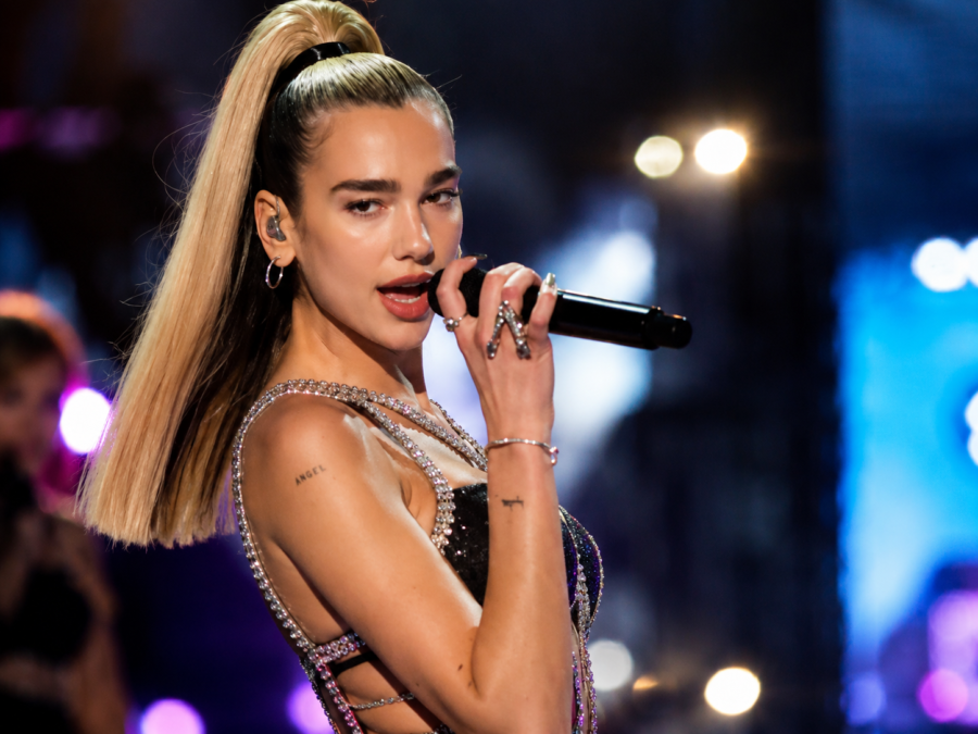 Dua Lipa: The British Pop Star Who Became a Global Fashion and Music Icon A stylish female pop singer performing live on stage, holding a microphone, with a high ponytail, sparkling black stage outfit, and dramatic concert lights in the background.